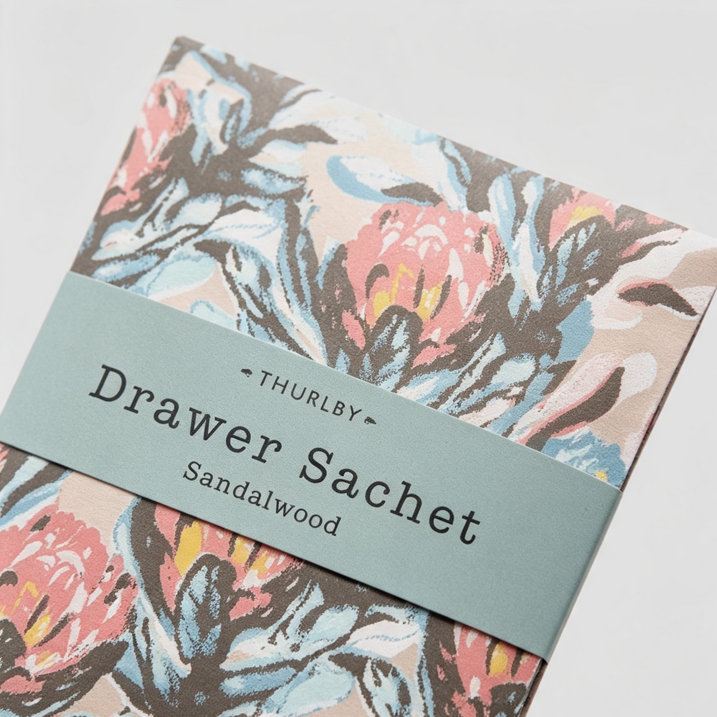 A close-up of a THURLBY Drawer Sachet Sandalwood, showing a floral patterned exterior with a dusty blue-green paper band wrapped around it.