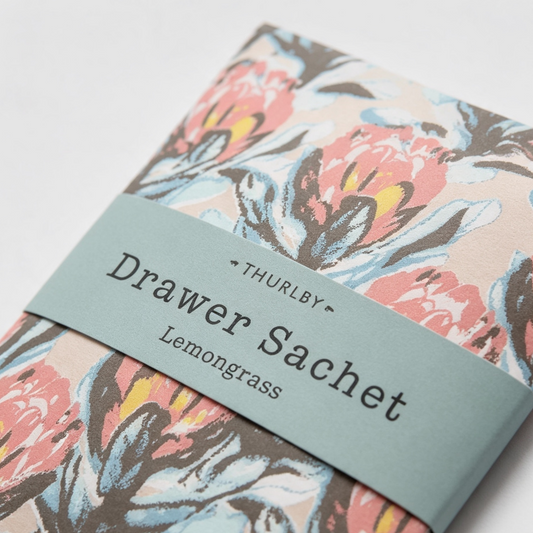 A close-up of a patterned fabric item wrapped with a dusty blue paper band labeled 'THURLBY Drawer Sachet Lemongrass.' The fabric has a floral print in pink, blue, brown, and cream tones on a light background.