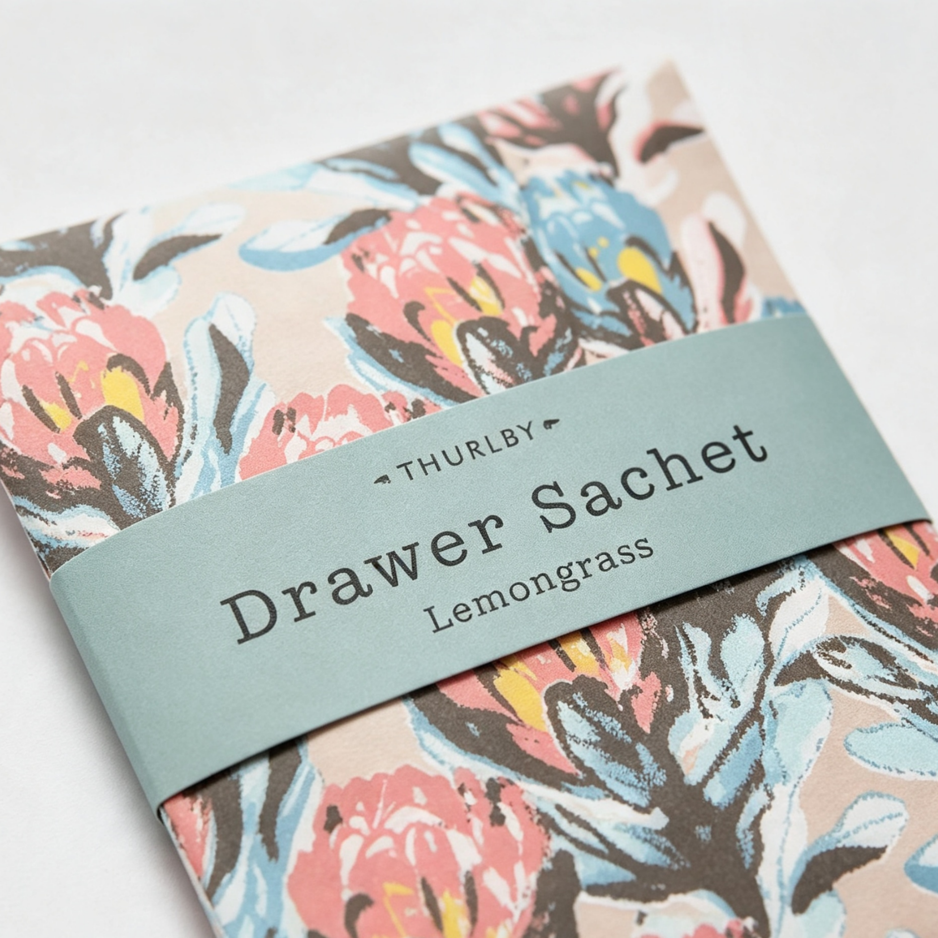 A folded paper product wrapped with a sage green paper band labeled 'THURLBY Drawer Sachet Lemongrass,' set against a floral patterned backing in pink, blue, beige, and brown tones.