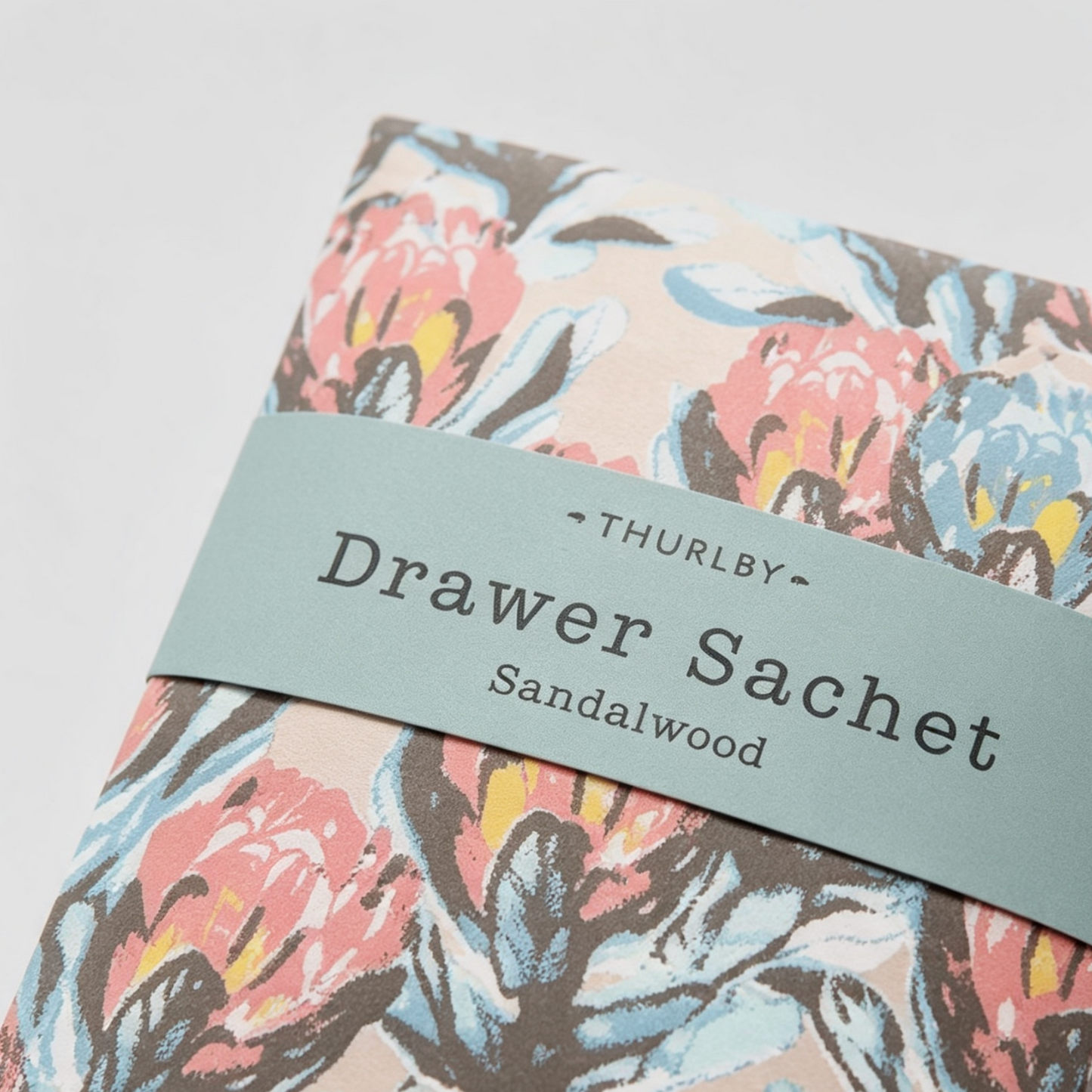 A close-up of a floral-patterned Drawer Sachet from Thurlby, featuring a dusty blue-green paper band wrapped around a fabric sachet with a coral, blue, and brown floral print on a light beige background.
