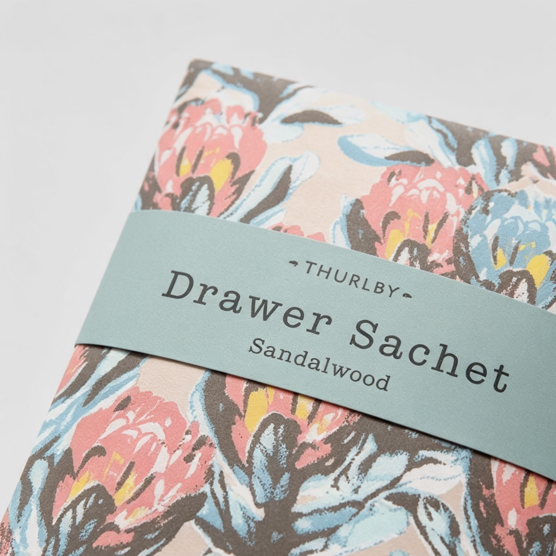 A close-up of a floral-patterned Drawer Sachet from Thurlby, featuring a dusty blue-green paper band wrapped around a fabric sachet with a coral, blue, and brown floral print on a light beige background.